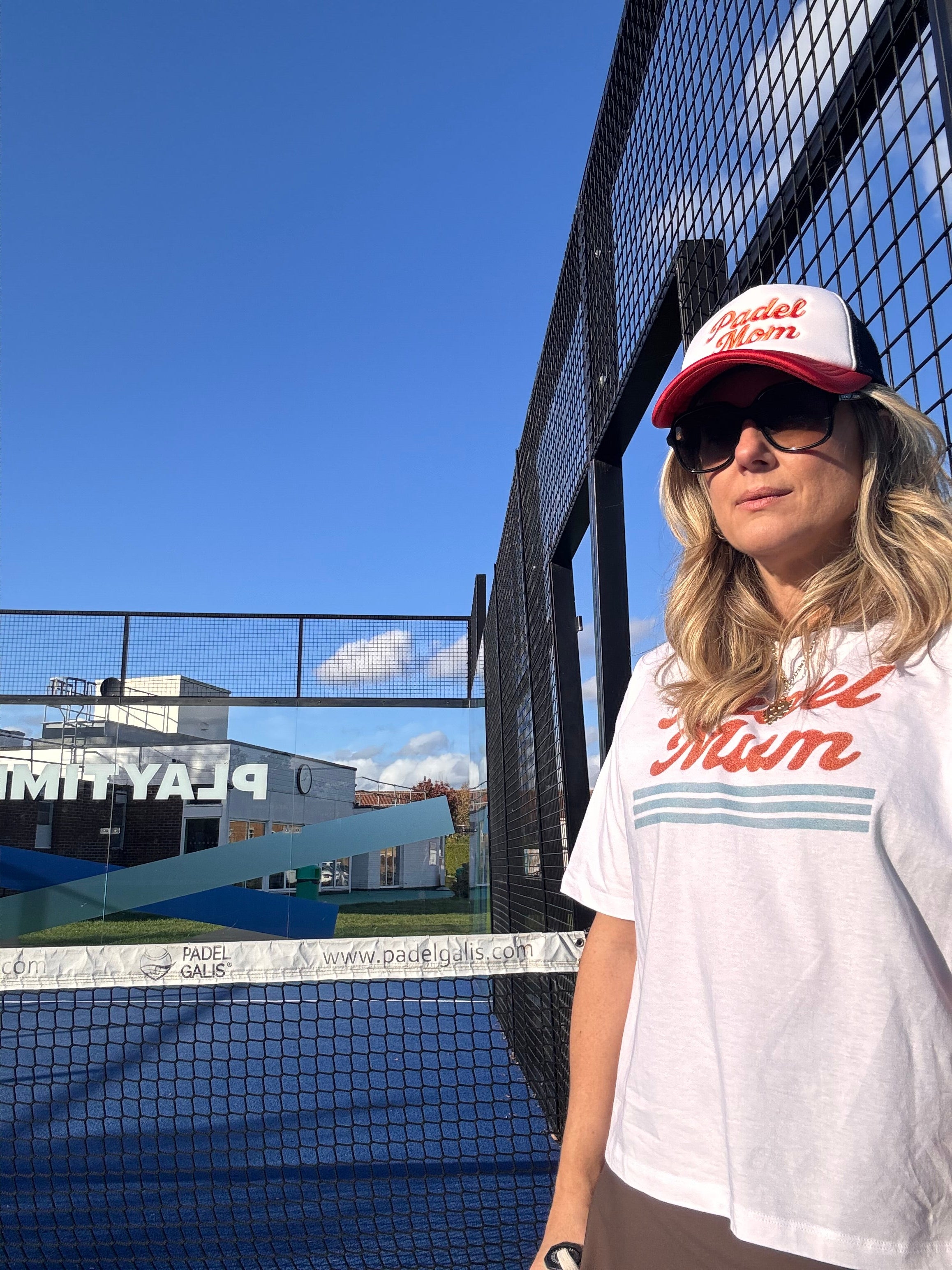 Person wearing a white t-shirt with red text and a red cap on a blue sky background on a padel court