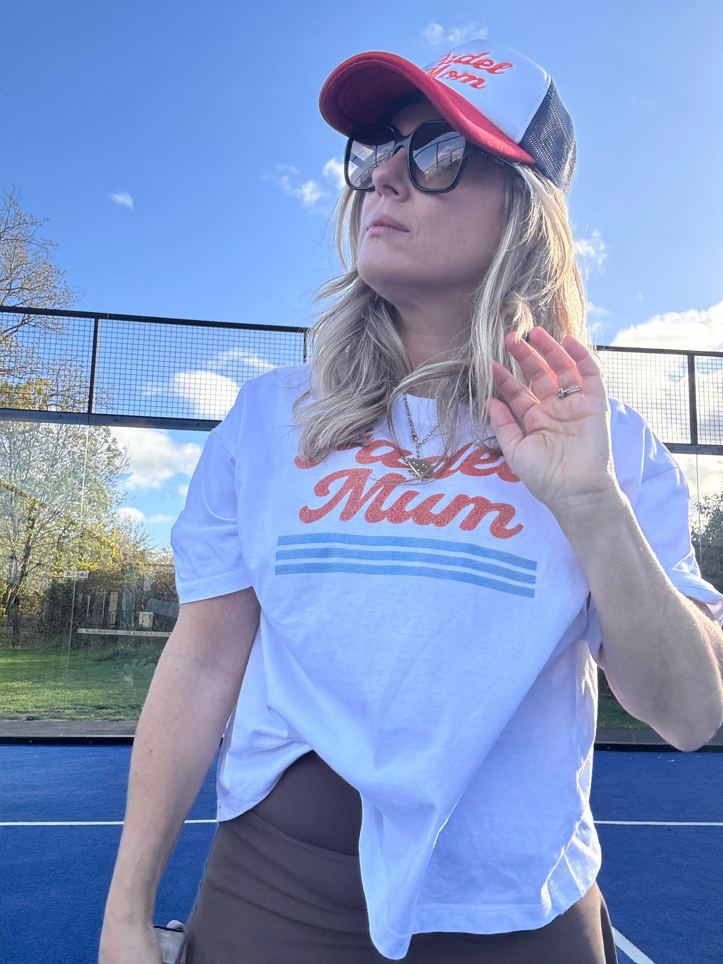 Person wearing a white t-shirt with ' Padel Mum' printed on it, standing outdoors.